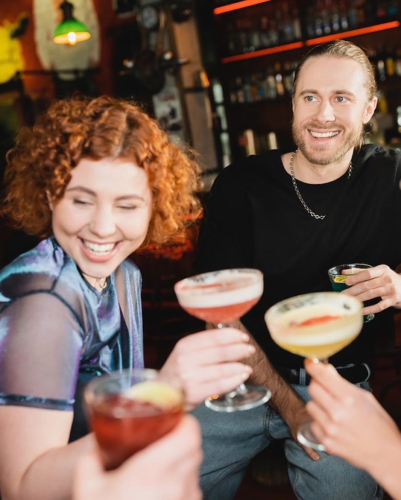 Positive man holding blue lagoon cocktail near interracial friends talking in bar